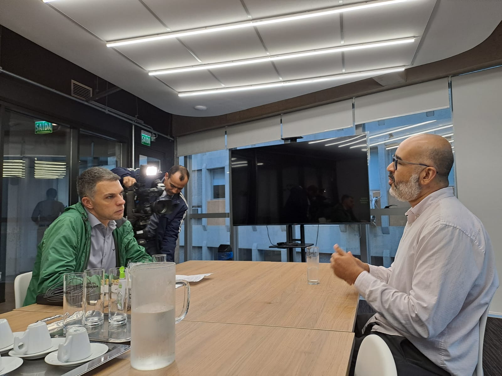 Tiago e entrevistador à mesa, com uma televisão ao fundo